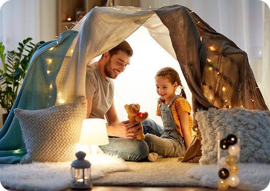 A father and daughter in a blanket fortress with lights.